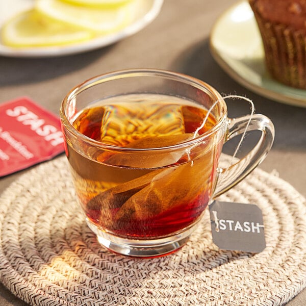 A glass cup of Stash English Breakfast tea with a tea bag in it on a table with a cupcake.