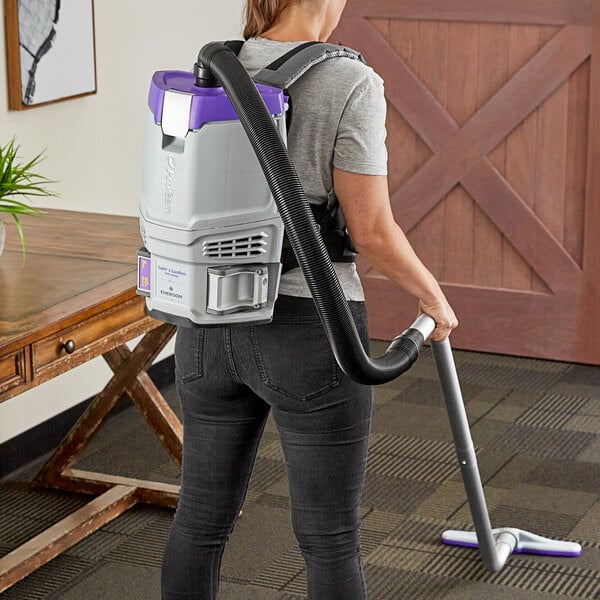 A woman using a purple ProTeam GoFit 3 cordless backpack vacuum to clean the floor.