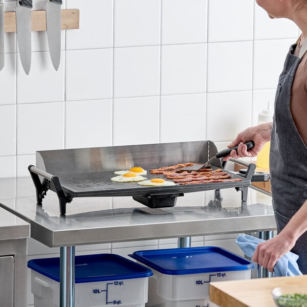 A light-duty electric countertop griddle being used to cook eggs and bacon in a commercial kitchen setting.