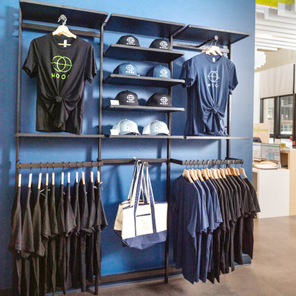 A UDIZINE black wall-mounted retail clothing display with faceouts and hanging rails holding black and blue shirts.