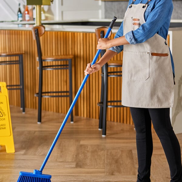 A woman in an apron using a blue Lavex metal threaded broom handle to clean a floor in a professional kitchen.