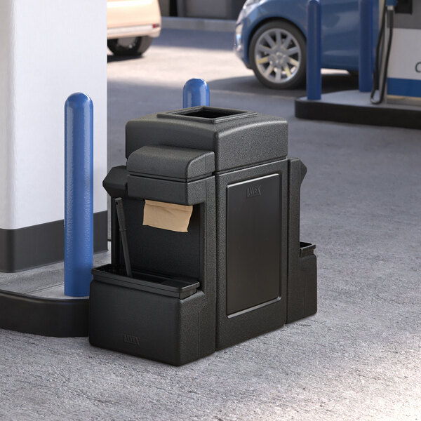 A black trash can with a brown bag inside sitting in front of a gas station.