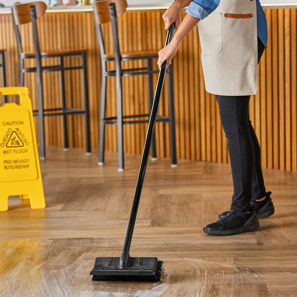A person using a Lavex black deck brush with a squeegee to clean a floor.
