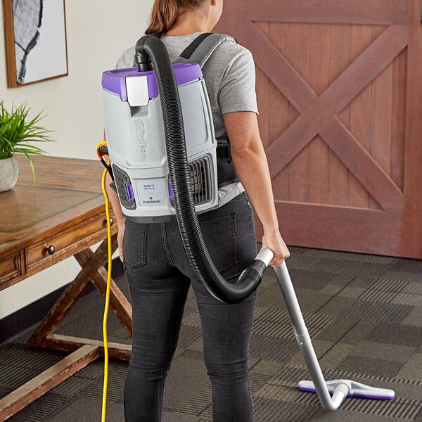 A woman using a ProTeam GoFit backpack vacuum with a purple and black tube to vacuum the floor.