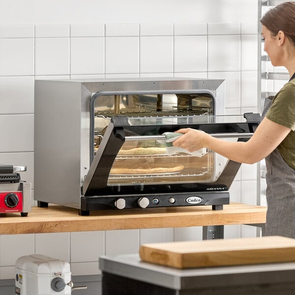 A stainless steel Cadco Arianna XAF-133 countertop convection oven with a glass door and manual controls, shown in use on a wooden counter.