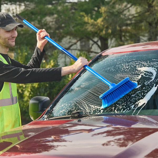 A blue Lavex Flo-Thru 10-inch floor and vehicle scrub brush with a 60-inch metal handle being used to clean a car windshield.