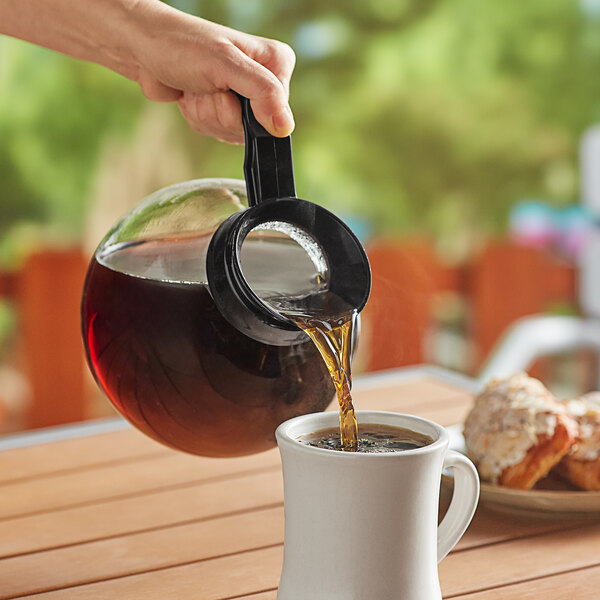 A person using a Curtis Crystalline glass coffee decanter with a black handle to pour coffee into a cup.