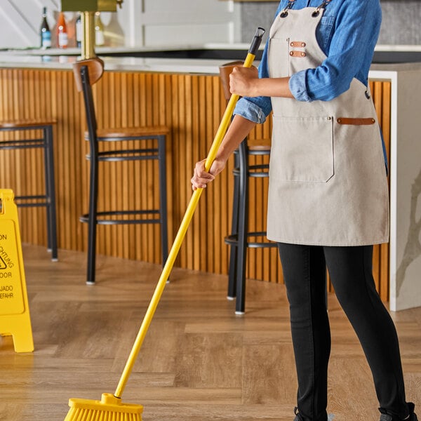 A woman in an apron sweeping a floor with a yellow Lavex metal broom handle.