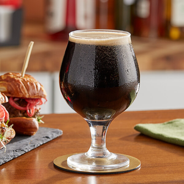 A glass of dark liquid on a coaster next to a sandwich on a table.