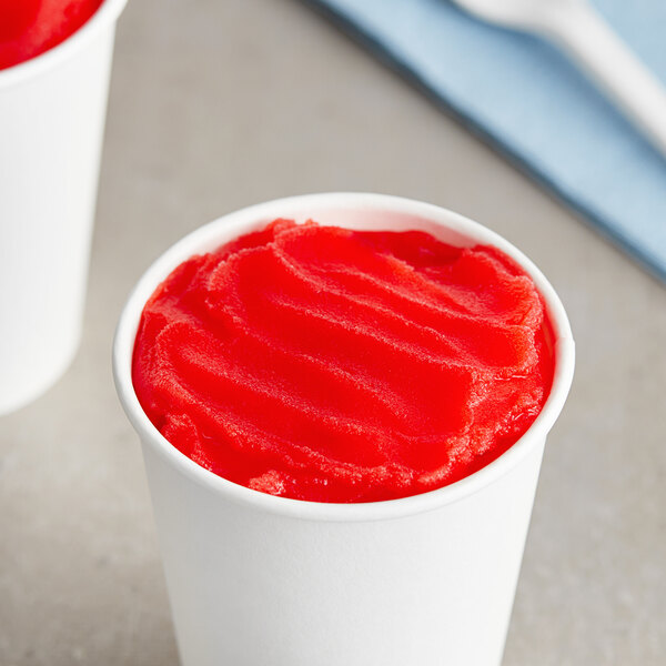 A white cup filled with red Tiger's Blood Italian ice next to a spoon and fork.