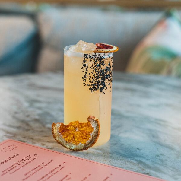 A tall glass filled with a light yellow beverage, ice, a dried citrus garnish, and a rim partially coated with black seeds, placed on a marble surface next to a dried citrus slice and a pink menu.