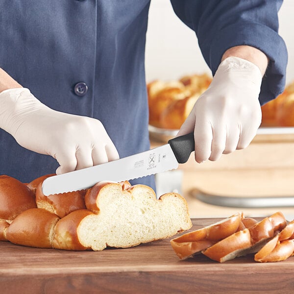 A person using a Mercer Culinary bread knife to cut a loaf of bread on a wooden cutting board.