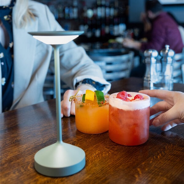 Two colorful cocktails in short glasses are placed on a wooden table next to a modern table lamp, with two people sitting nearby.