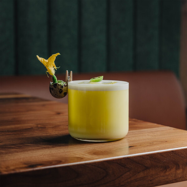 A yellow cocktail with a frothy top is served in a clear glass, garnished with a small flower and a decorative element clipped to the rim, placed on a wooden table.