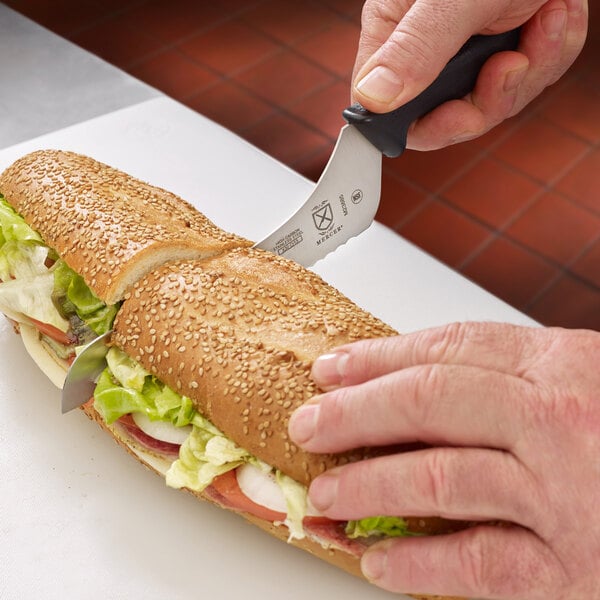 A hand using a Mercer Culinary Millennia offset serrated knife to cut a sandwich.
