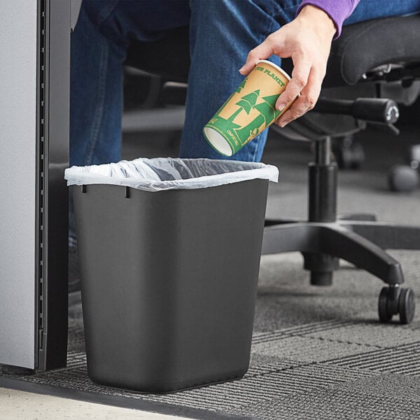 A person holding a paper cup throws it into a Lavex black rectangular trash can.