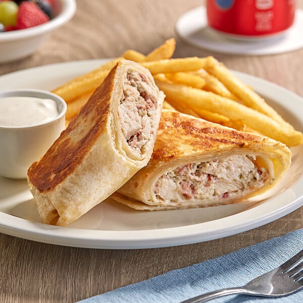 A red and white Clara's Kitchen Chicken Bacon Ranch Wrap on a plate with fries and a cup of white sauce.