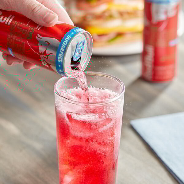 A hand pouring Red Bull Sugar-Free Watermelon Energy Drink into a glass.
