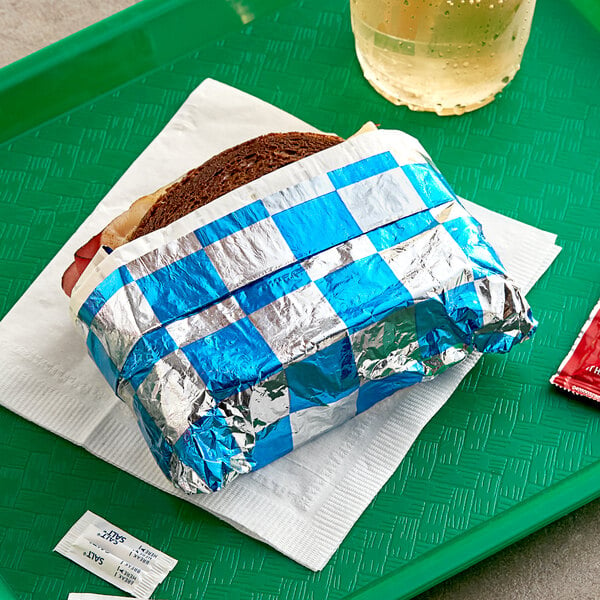 A green tray with a sandwich wrapped in Bagcraft blue and silver checker insulated foil and a glass of liquid.