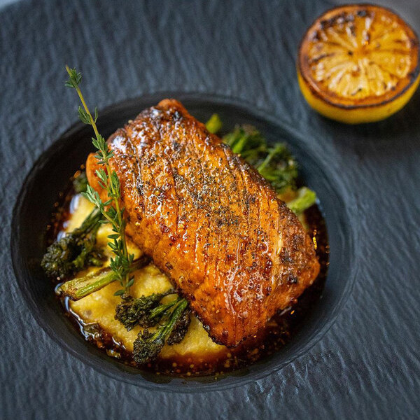 A vegan salmon filet served on a bed of vegetables and sauce, garnished with herbs and accompanied by a grilled lemon half.
