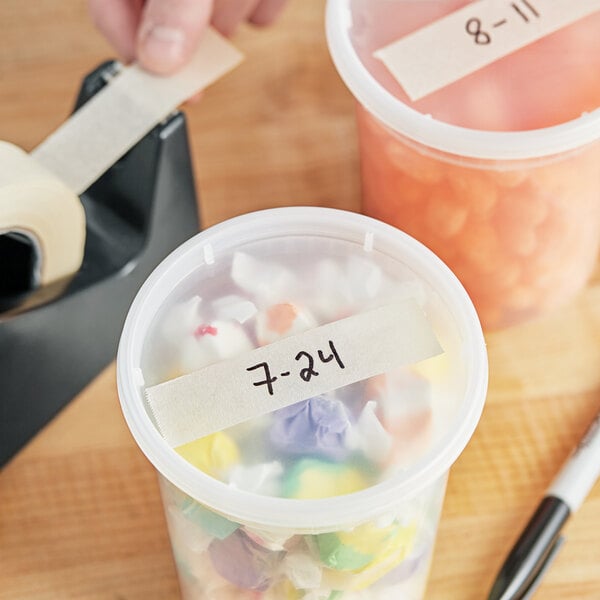 A tape dispenser on a table with a plastic container of candy and a white piece of paper with black text.