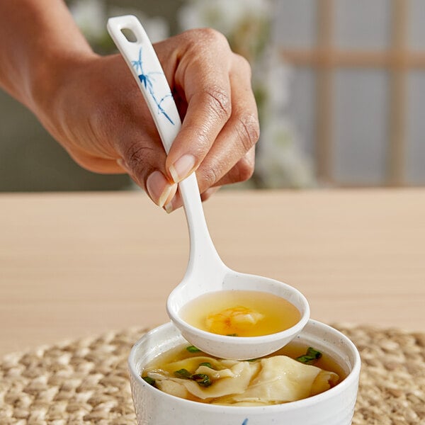 A hand holding an Acopa Biru white melamine soup ladle over a bowl of soup.