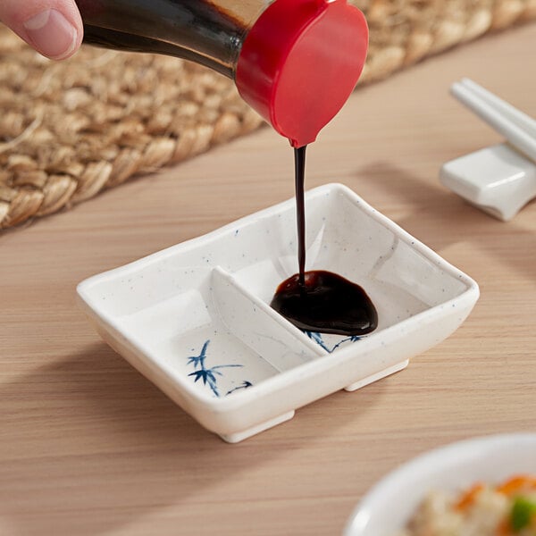 A hand pouring soy sauce into an Acopa Bambu Biru rectangular divided sauce dish.