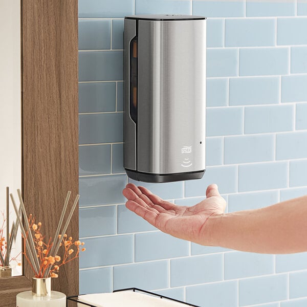 A person using a Tork stainless steel wall-mount automatic foam hand soap/sanitizer dispenser.