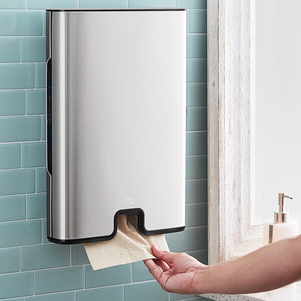 A hand using a Tork stainless steel wall-mount paper towel dispenser.