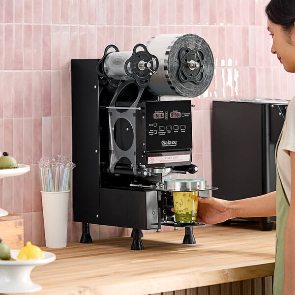 A woman using a Galaxy Automatic Cup Sealing Machine to seal a drink.