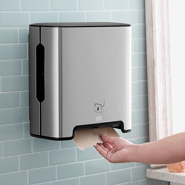 A hand reaching out to a Tork stainless steel wall-mount paper towel dispenser.