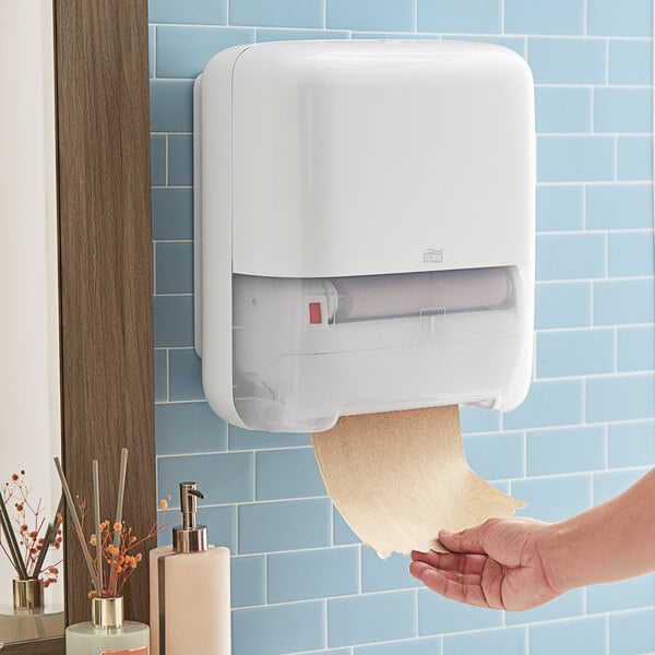 A hand using a Tork white plastic wall-mount paper towel dispenser with a roll of paper towels.