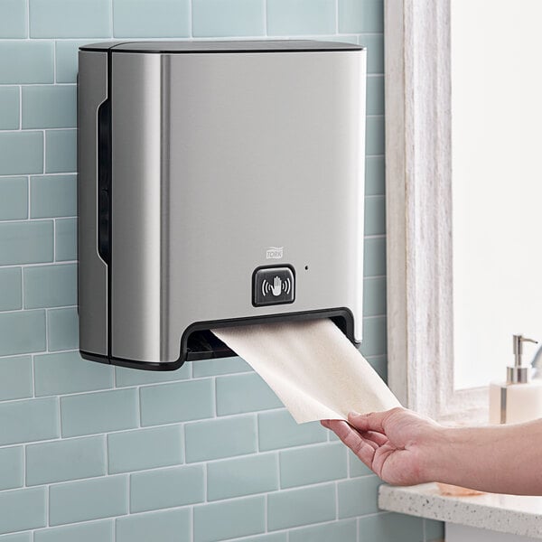 A hand using a Tork stainless steel wall-mount automatic paper towel dispenser.