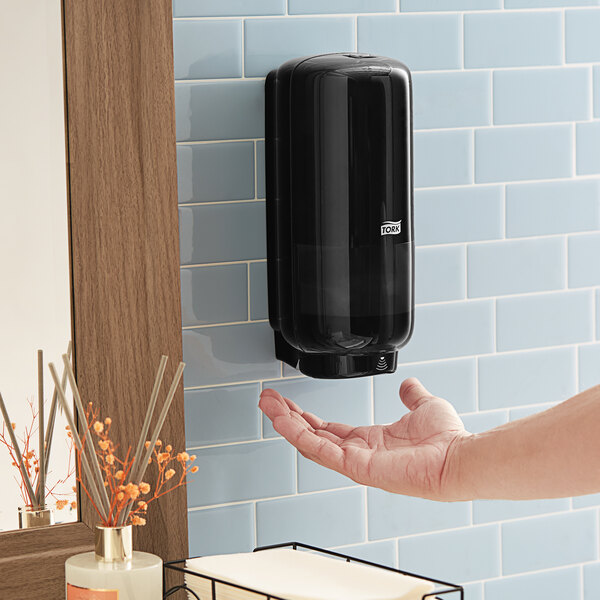 A person using a Tork black wall-mount automatic foam hand soap dispenser.