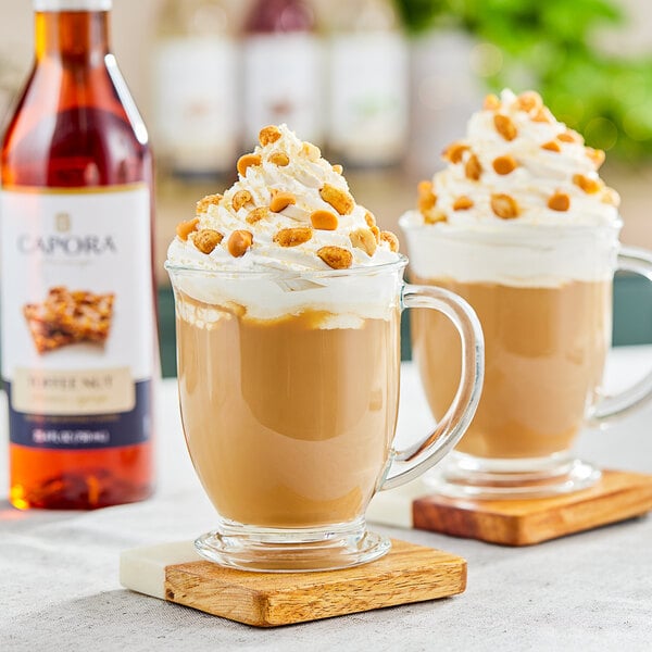 A bottle of Capora Toffee Nut flavoring syrup with a glass mug of coffee with whipped cream and nuts.