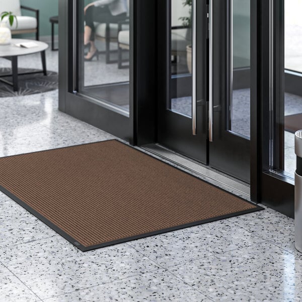 A brown Lavex Needle Rib entrance mat on a marble floor in front of a glass door.