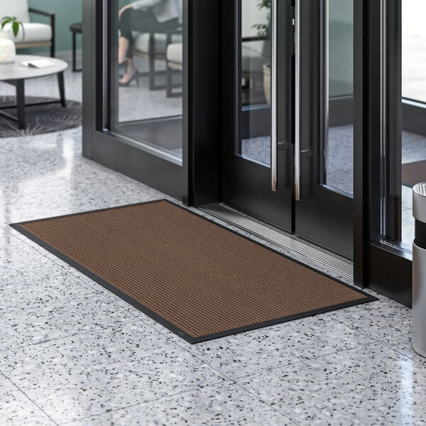 A brown Lavex Needle Rib entrance mat in front of glass doors.
