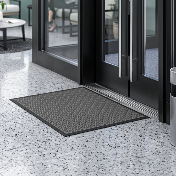 A gray Lavex parquet entrance mat on a white marble floor in front of a glass door.