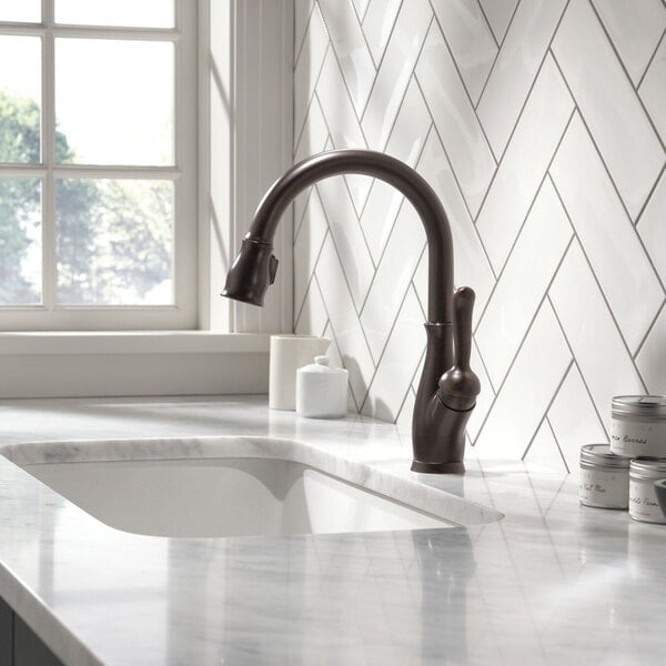 A white tile counter with a Delta Leland Venetian Bronze kitchen faucet above a sink.