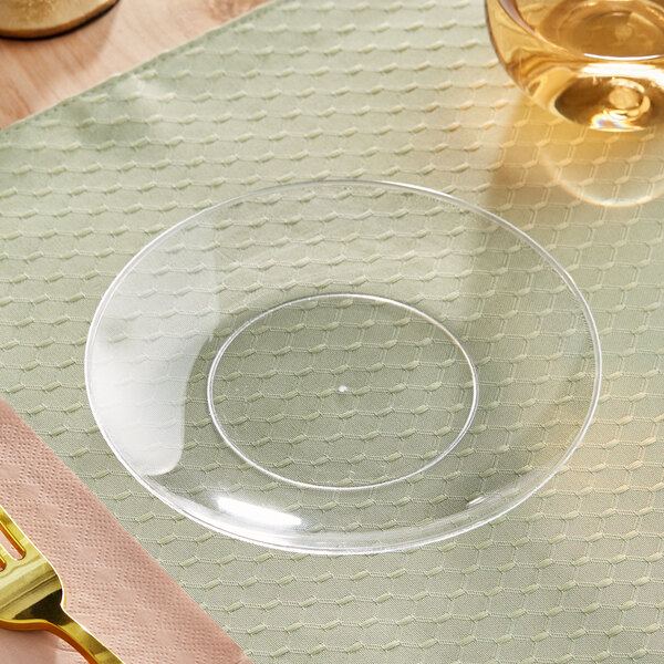 A clear Visions plastic coupe plate on a table with a glass of liquid.