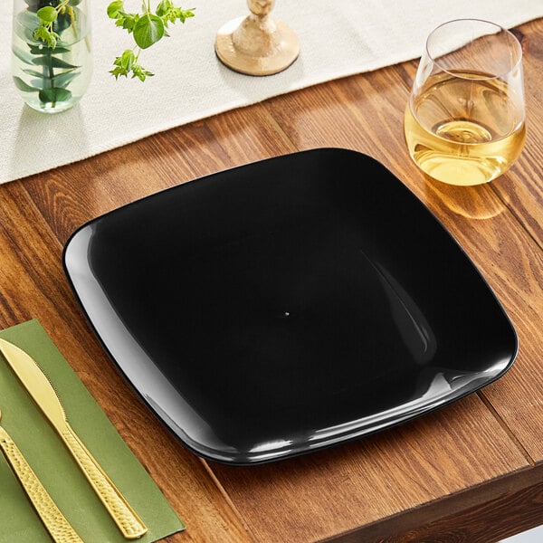 A black square Visions plastic coupe plate with a knife and fork on a wood table.