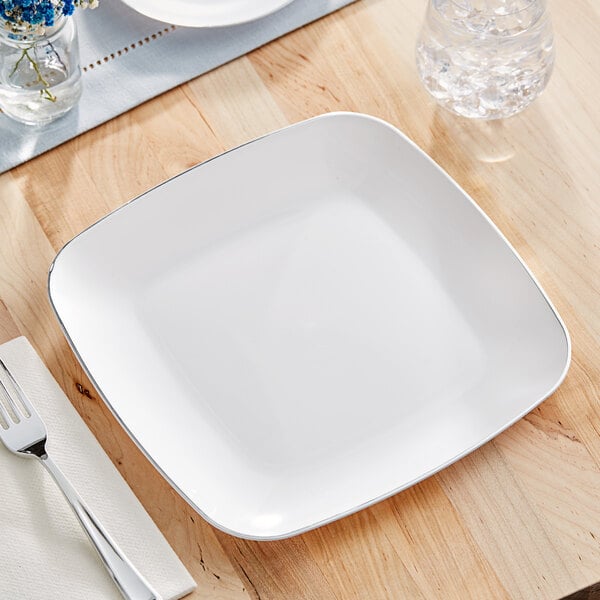 A white square Visions plastic plate with a silver fork and knife on a wood surface.