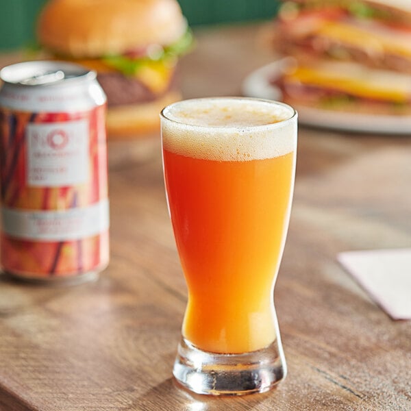 A glass of Untitled Art Mango Dragonfruit fruited sour juice next to a can of beer and a burger.