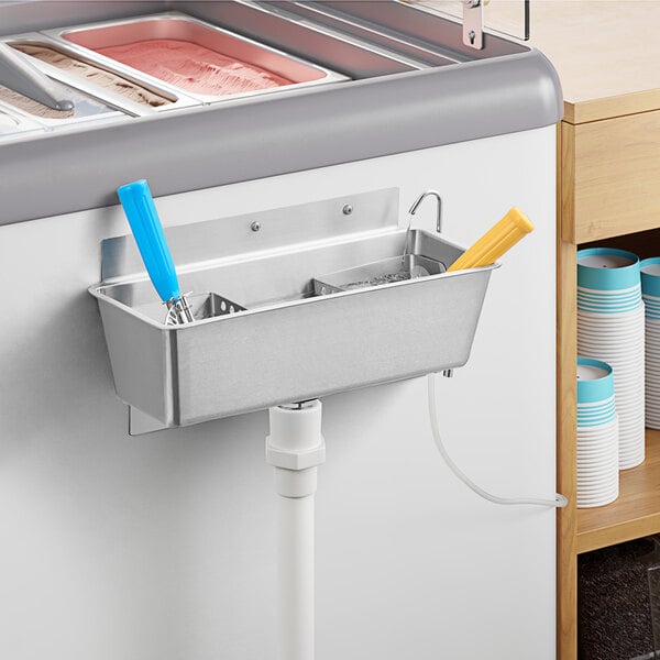 A metal container with a Regency blue handle attached to a white pipe over a metal sink with two scoops in it.