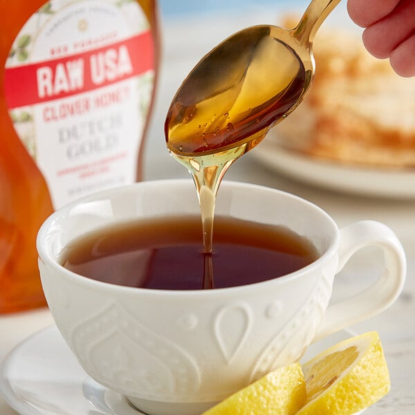 A spoon of Dutch Gold Raw Clover Honey being poured into a cup of tea with a lemon wedge.