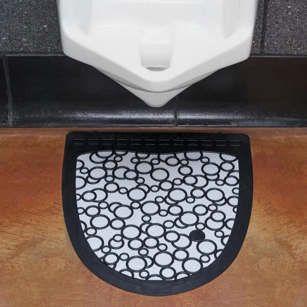 A black and white disposable urinal floor mat placed in front of a urinal.