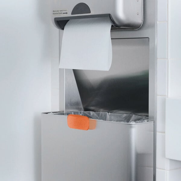 An orange air freshener clip attached to a stainless steel trash bin below a paper towel dispenser in a restroom.