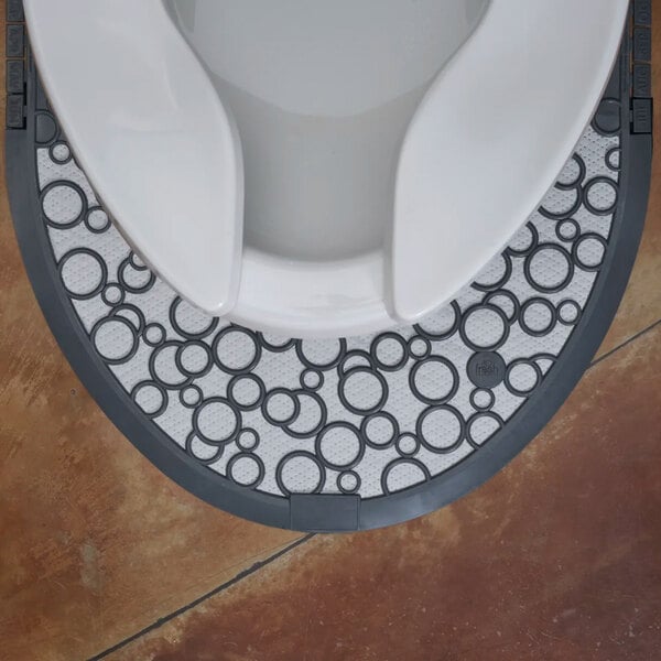 A gray and white disposable toilet floor mat with a circular pattern placed around the base of a toilet.