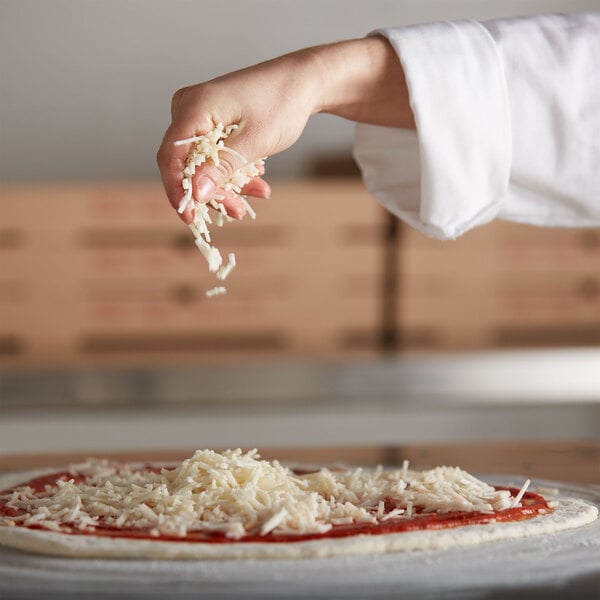 A person's hand sprinkling Daiya vegan shredded mozzarella cheese on a pizza.