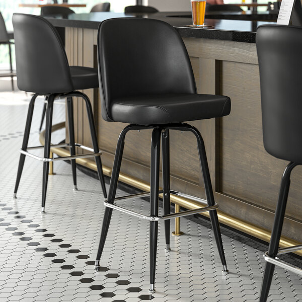 Three Lancaster Table & Seating black vinyl bucket seat cushions on black bar stools with metal legs at a bar counter.